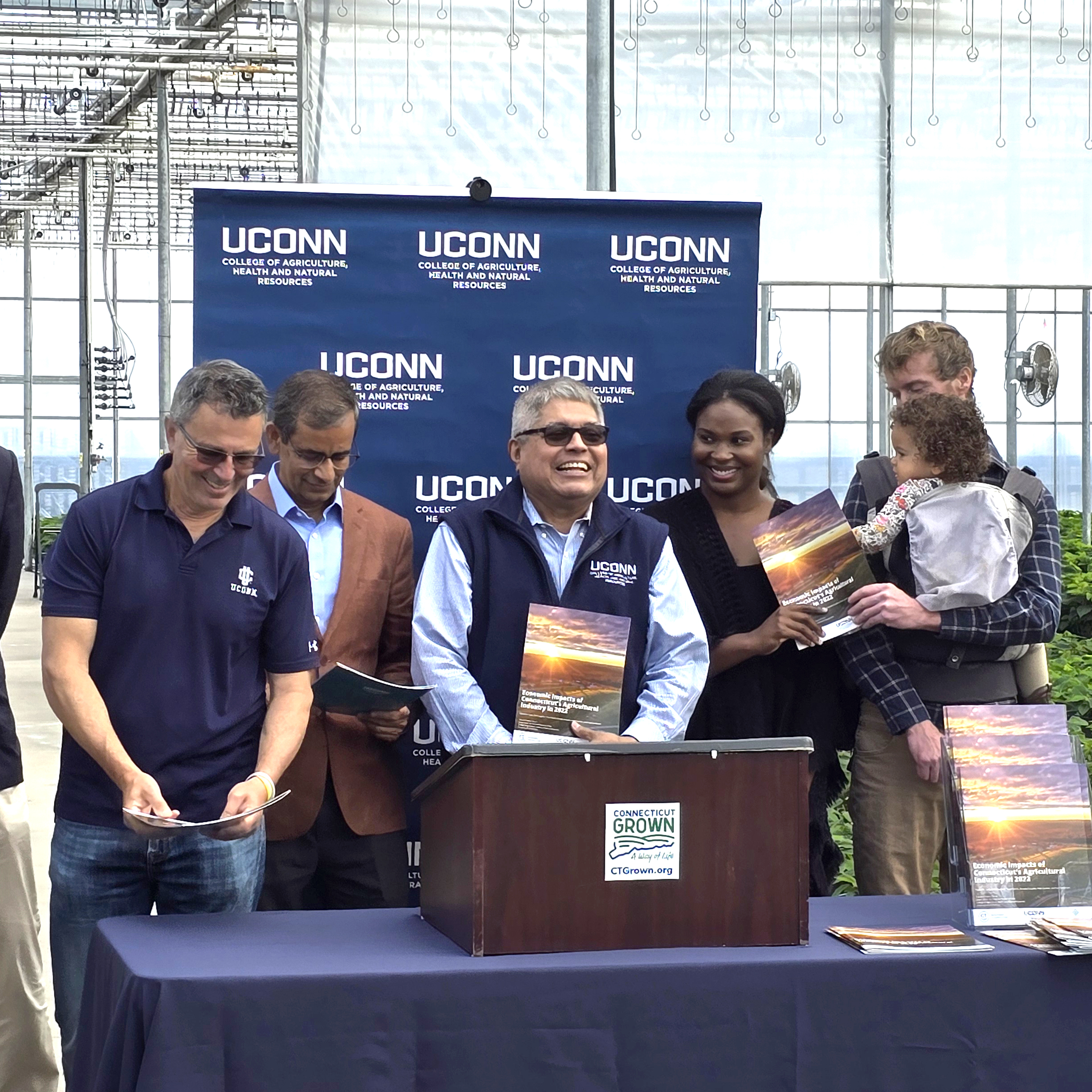 5 adults, one holding a baby, standing behind a podium in a greenhouse